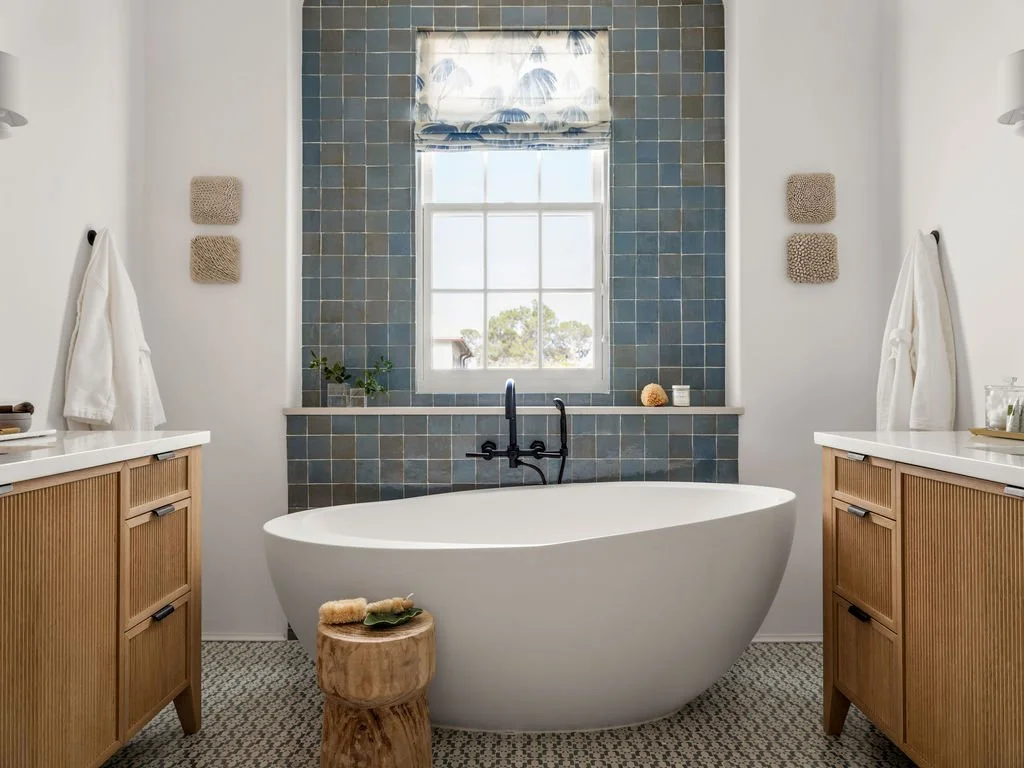 Modern bathroom with a white freestanding bathtub centered in front of a window with blue tile wall behind it, wooden vanities on each side, and neutral decor.