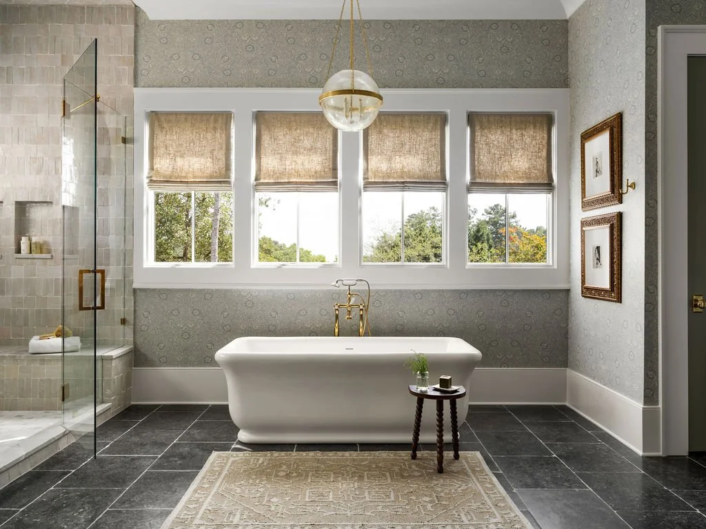 A bathroom featuring a freestanding bathtub in front of four windows, a small wooden side table with a plant, framed artwork on the wall, and a glass-enclosed shower on the left.