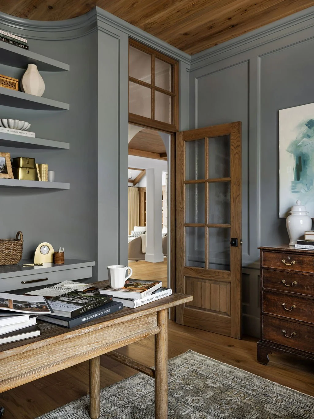 Interior view of a cozy living space with a wooden table, open books, a mug, and decorative items on a sideboard, with a door leading to a bright room in the background.