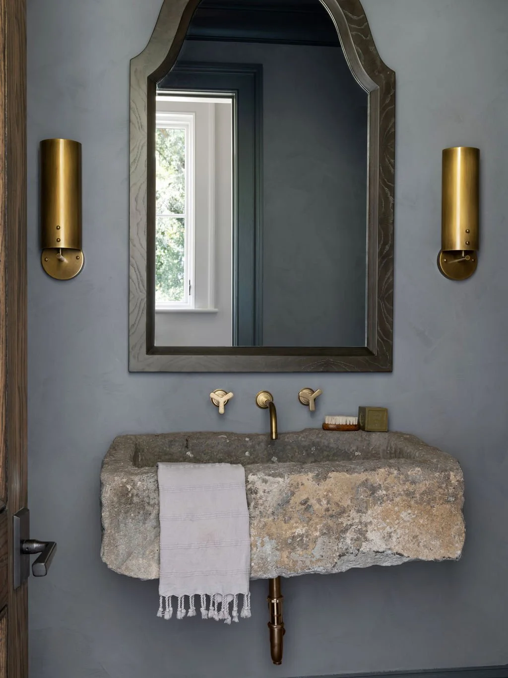 Bathroom with a rustic stone sink, gold fixtures, a large mirror framed with wood, and a window reflecting greenery outside.