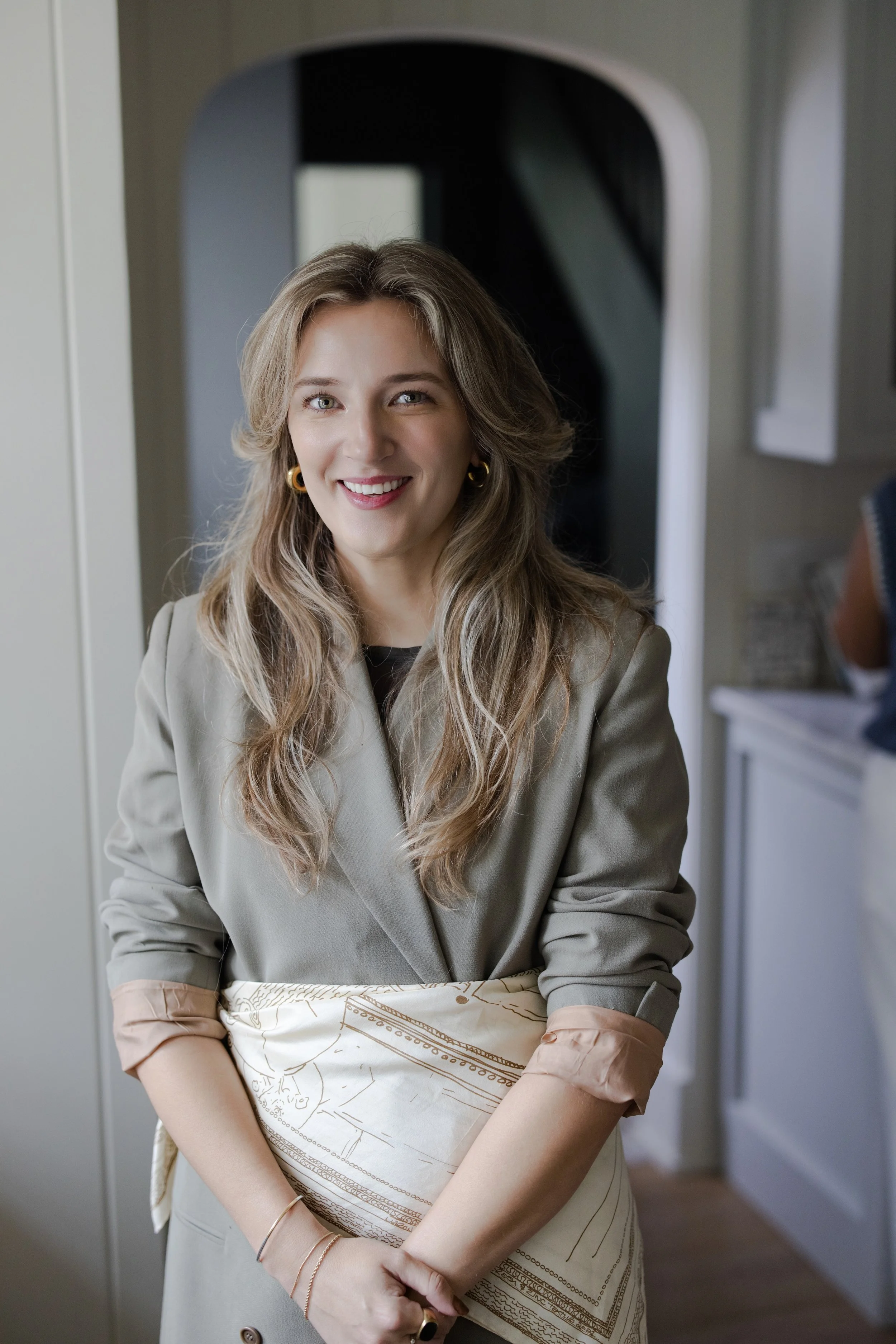 A young woman with long wavy blonde hair, wearing a gray blazer and an apron, smiling in a kitchen setting.