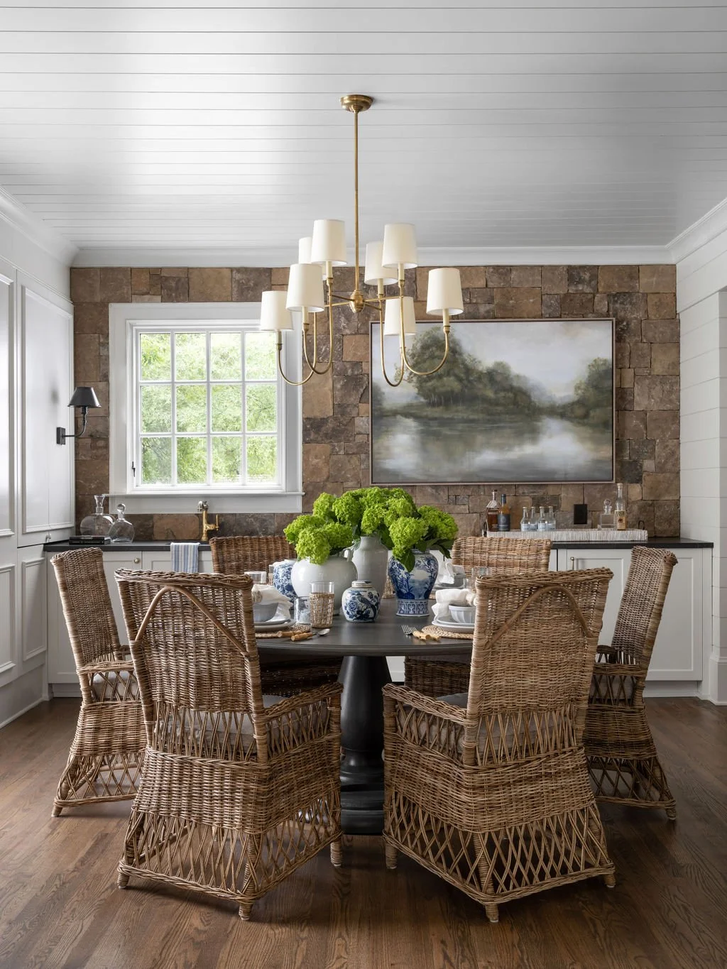 A dining room with a round black table surrounded by eight wicker chairs, set with white plates, napkins, and glasses. A large white vase filled with green flowers is in the center of the table. The room has a window with white trim, a stone accent w