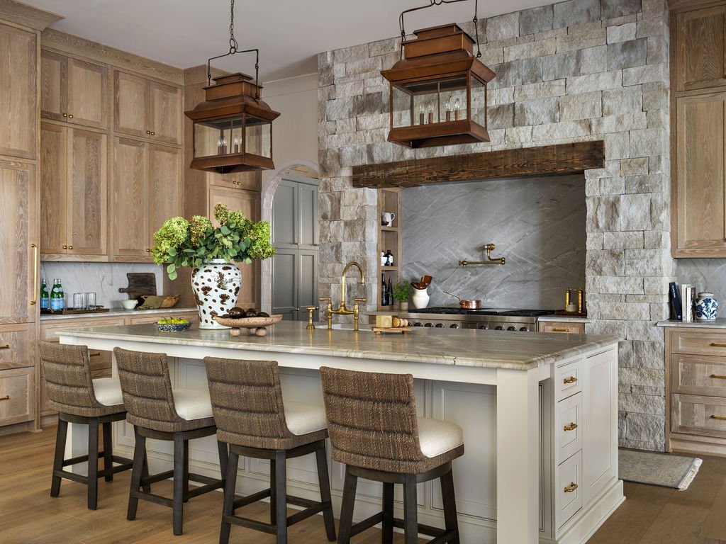 Modern kitchen with light wood cabinets, a white island with a vase of green flowers, five chairs, stone wall, wooden beams, hanging lanterns, and a stainless steel stove.