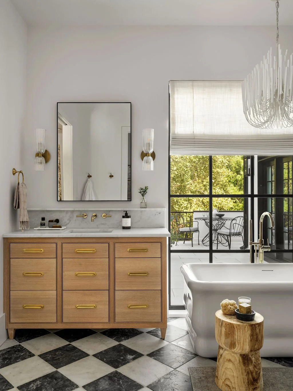 A modern bathroom with a wooden vanity, white sink, gold fixtures, a large mirror, wall-mounted lights, a freestanding bathtub, and a view of an outdoor patio with a table and chairs through a sliding glass door.