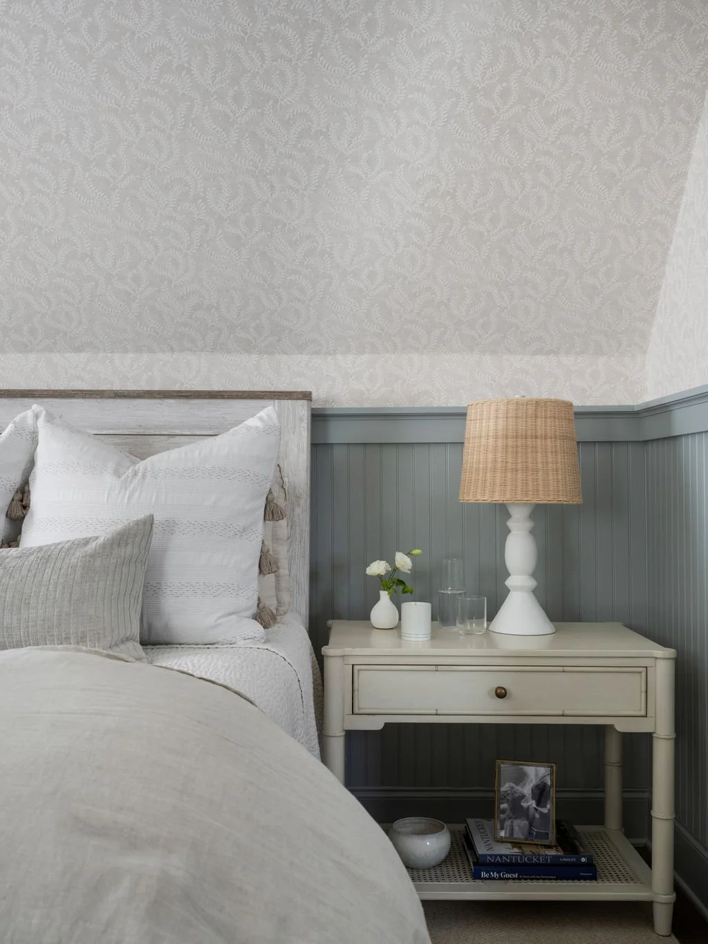 A cozy bedroom corner with a white bed featuring decorative pillows, a nightstand with a beige lamp, a small vase with white flowers, and books, set against a half wall with paneled wainscoting and patterned wallpaper.