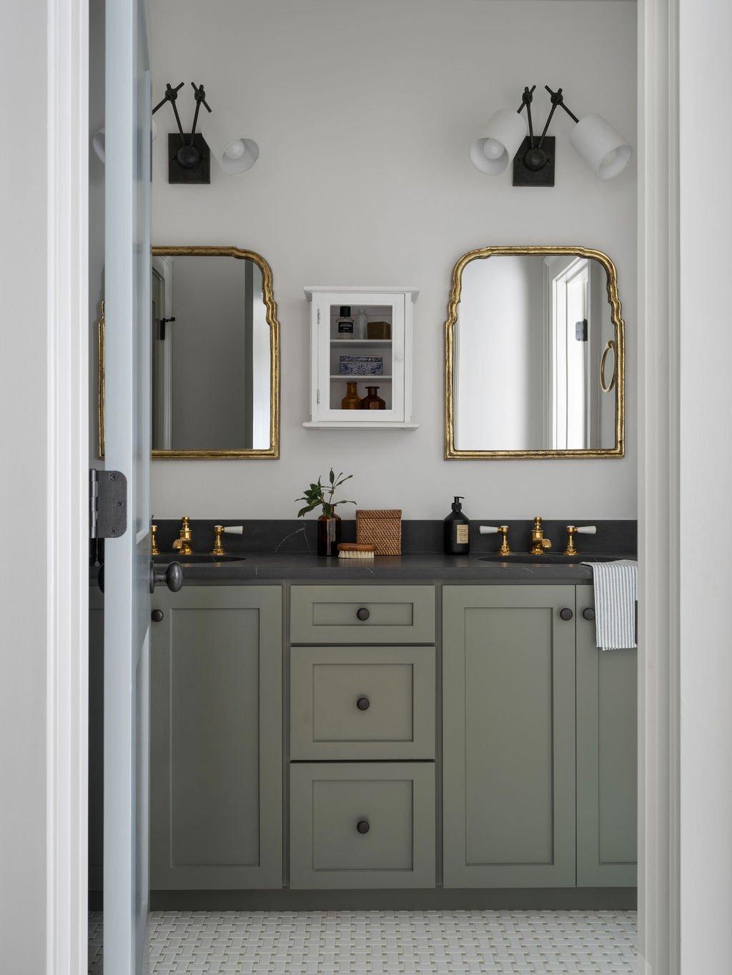 Bathroom with gray cabinets, black countertop, two mirrors with gold frames, black and white light fixtures, small white wall cabinet, and minimal decor.