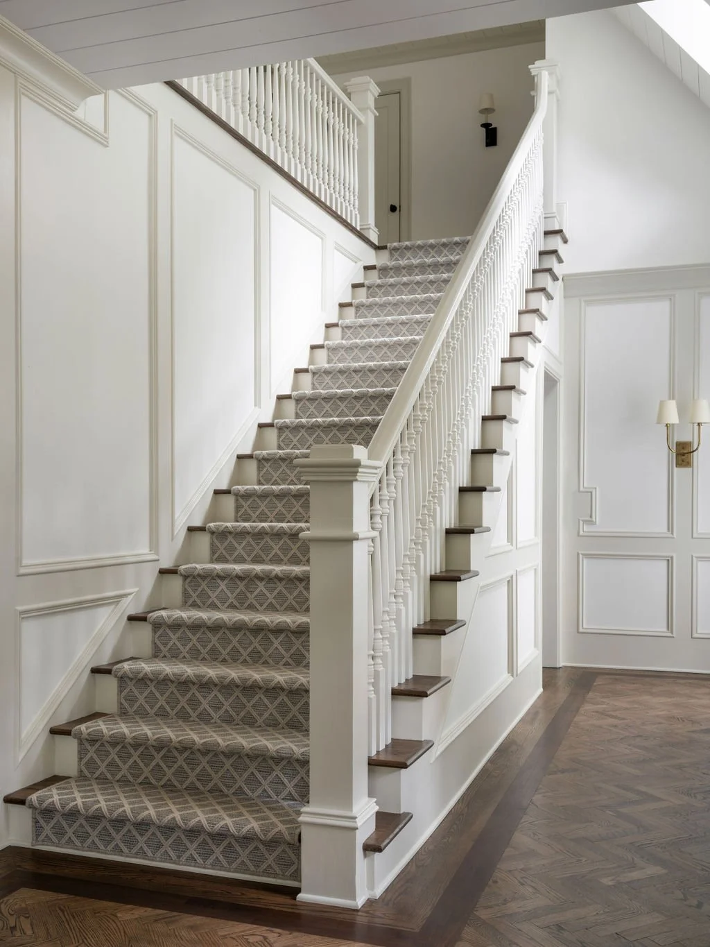 Interior staircase with beige carpet and wooden steps in a white-walled home, featuring decorative wall paneling and a wall-mounted lamp.