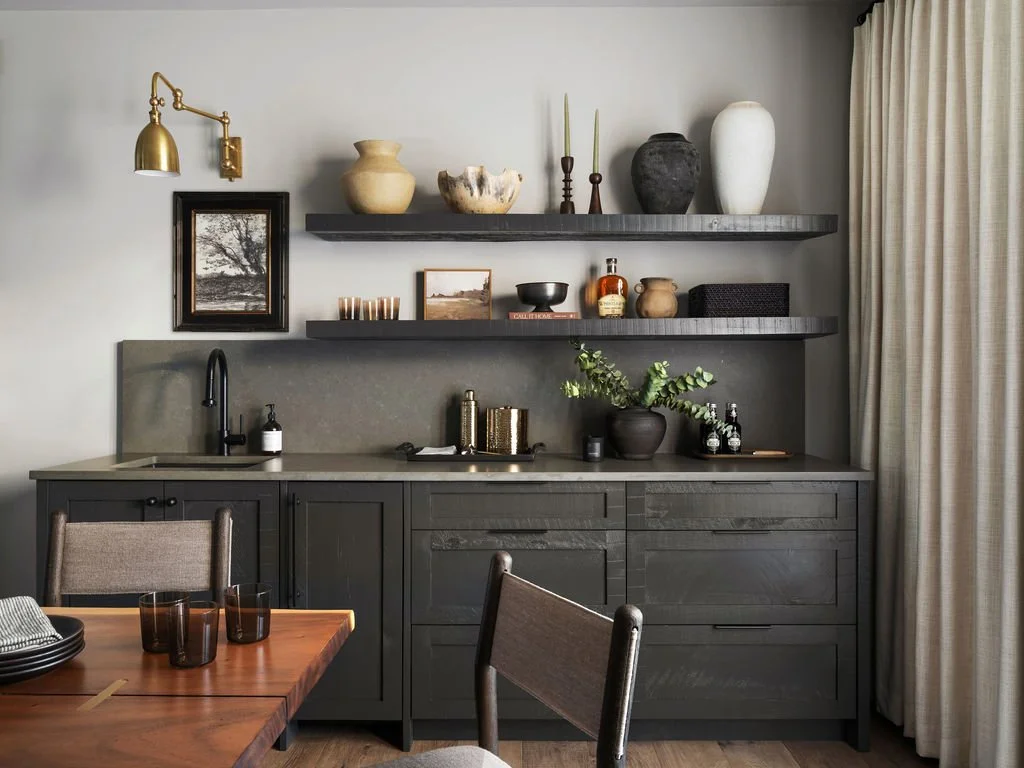 A modern kitchen with dark gray cabinets, a gray countertop, and floating shelves holding decorative vases, candles, and small decorative objects. There is a small black sink with a black faucet, a black soap dispenser, and a potted plant. To the rig