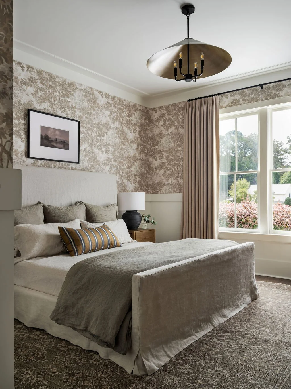A bedroom with a large window showing trees outside, a bed with beige bedding and multiple pillows, a nightstand with a lamp, and beige curtains, in a cozy and elegant decor style.
