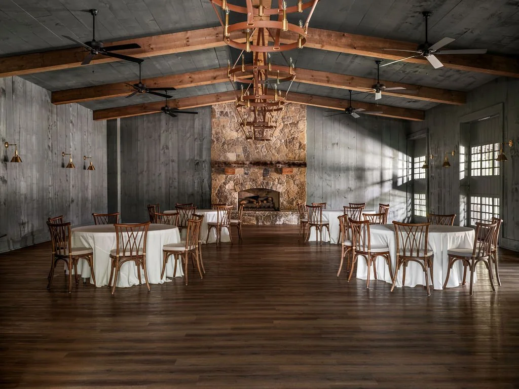 A spacious event hall with wooden floors and a large stone fireplace centered on the back wall. There are four round tables draped with white tablecloths and surrounded by wooden chairs. The ceiling has exposed wooden beams with hanging fans and chan