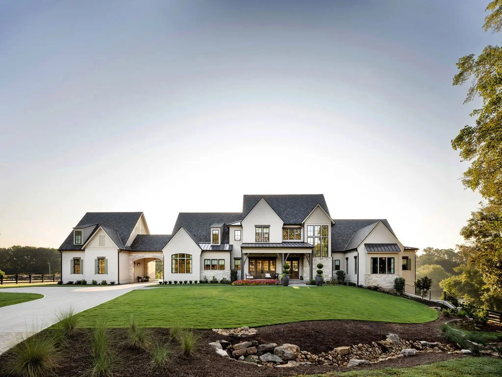 Large modern house with white exterior, multiple gabled roofs, and large windows, surrounded by a well-manicured lawn and trees, under a clear sky.