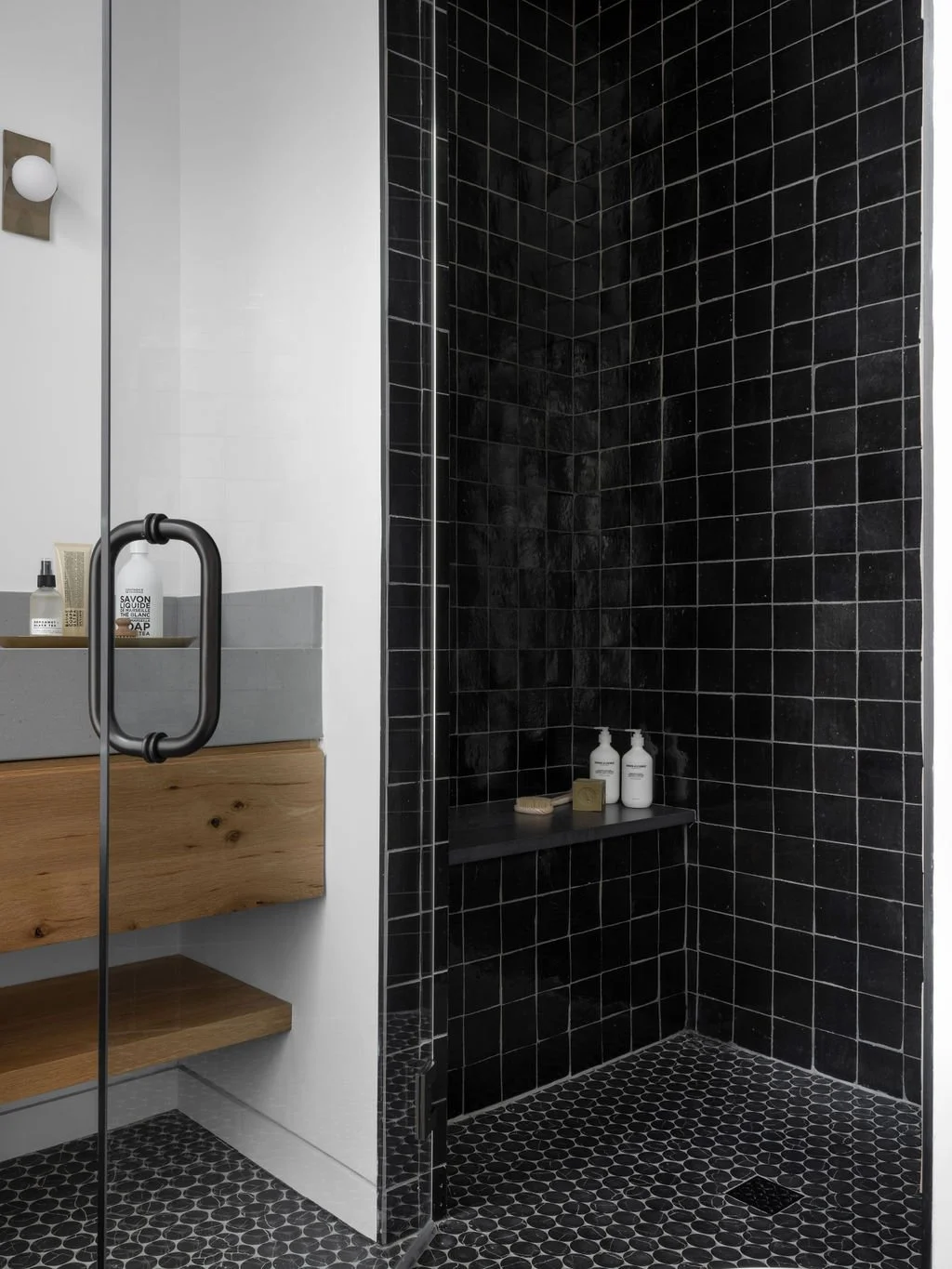 Modern shower area with black tiled walls and floor, a black bench with bottles of toiletries, and a glass door with a black handle.