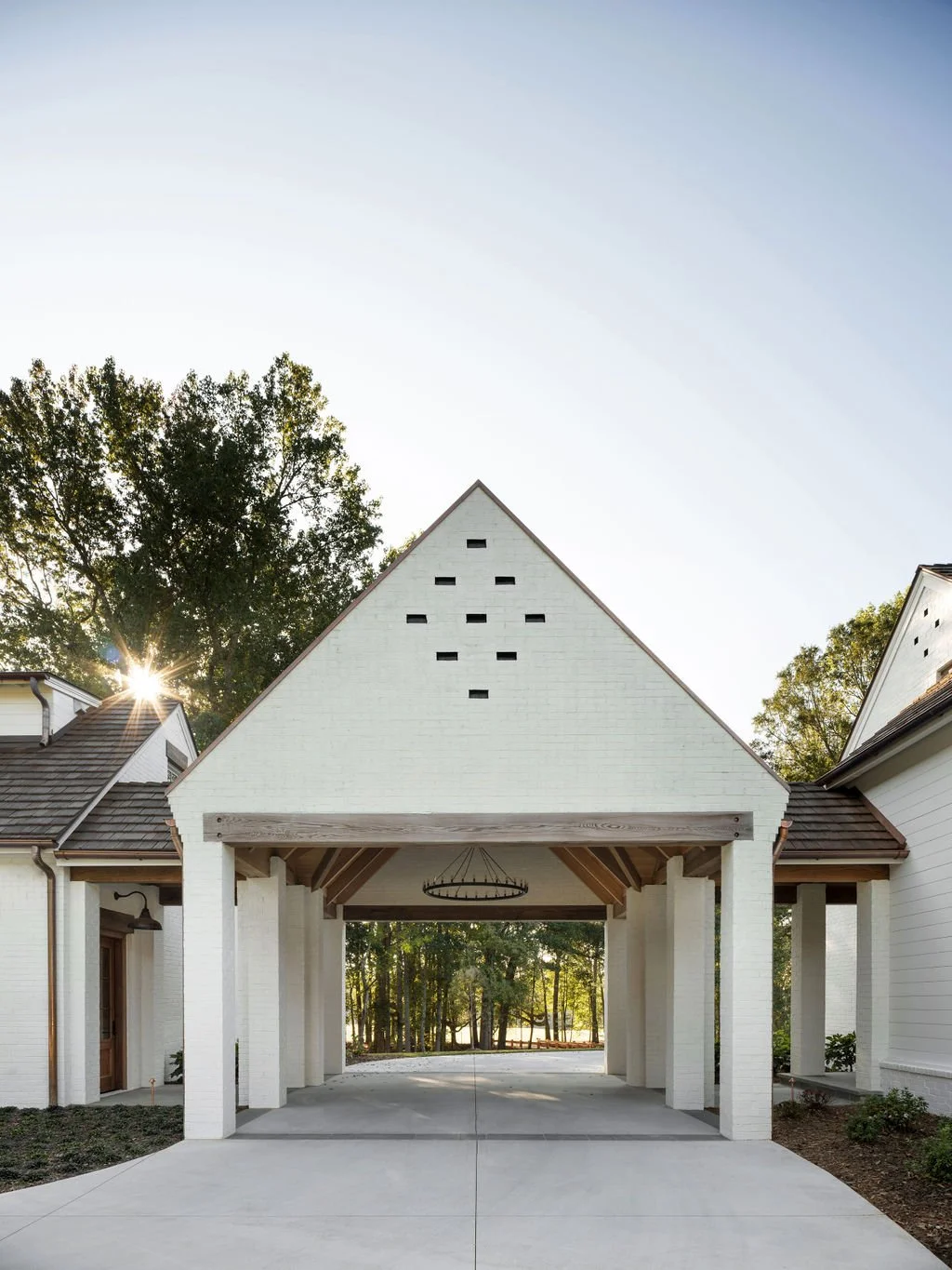 A white house with an open carport, triangular roof, and decorative black vent blocks, set against trees with the sun shining in the background.