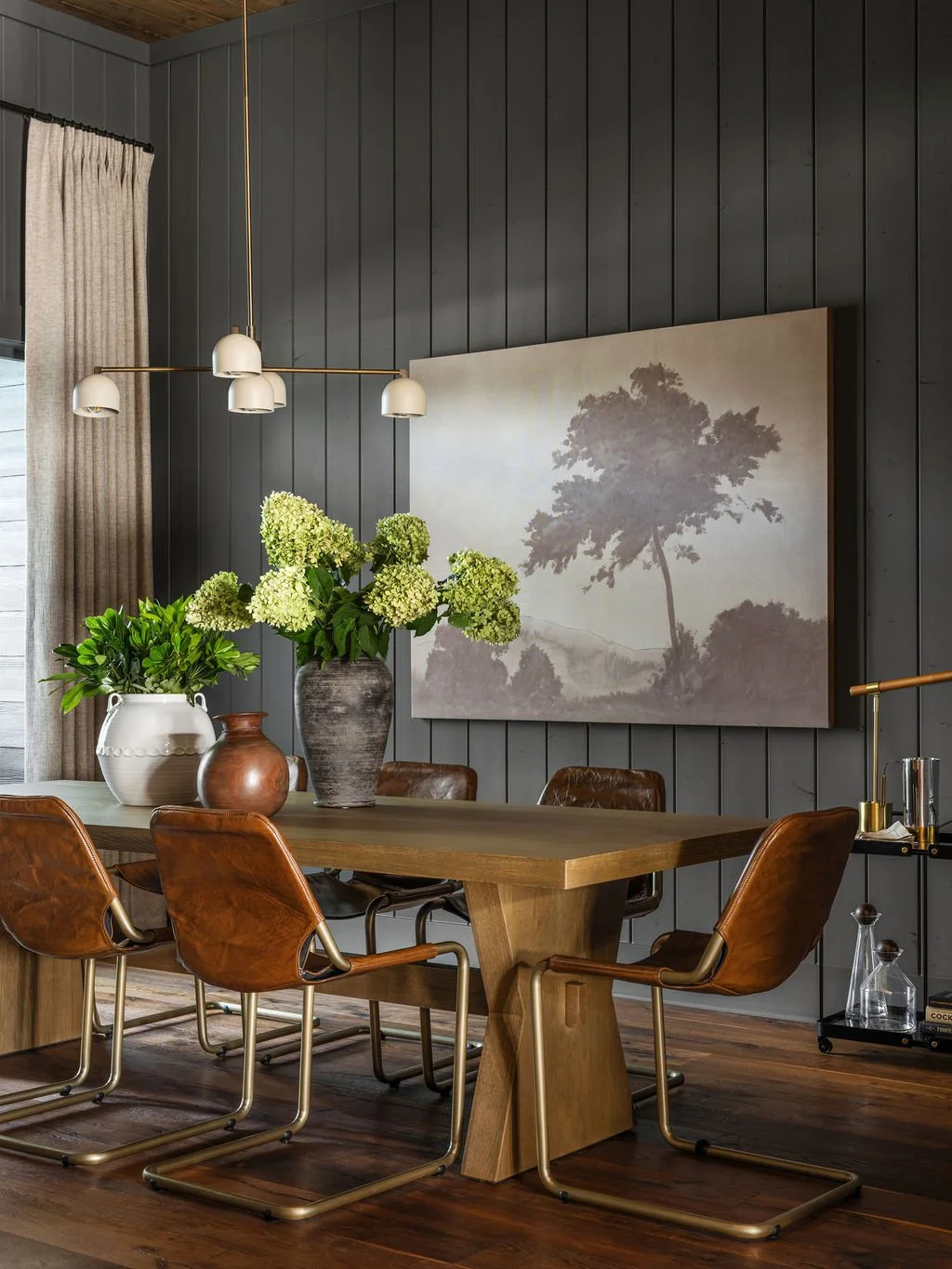 A dining room with a wooden table and five leather chairs. On the table are three vases with green hydrangeas and foliage. Behind is a dark paneled wall with a large landscape painting of a tree. A modern chandelier with five white shades hangs above the table. To the left, a window with beige curtains is partially visible, and to the right, a bar cart with glassware and a gold lamp.