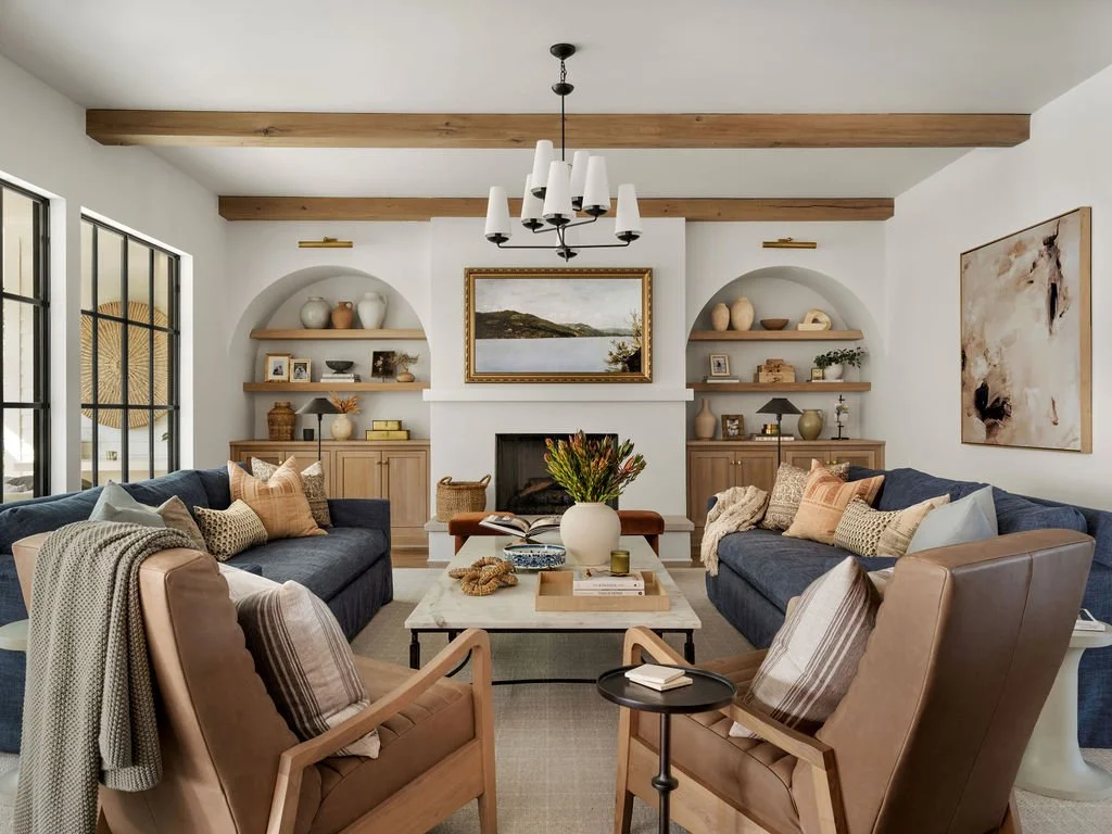 A living room with two navy sofas, two tan armchairs, a white fireplace with a landscape painting, wooden shelves with decorative items, a chandelier, a coffee table with a vase of flowers, and large windows.