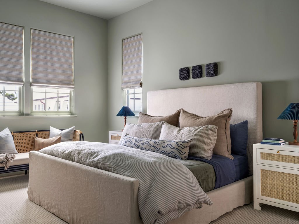 A bedroom with a large upholstered bed, multiple pillows, two nightstands with blue lamps, a sitting area with a wicker sofa, and windows with striped blinds.