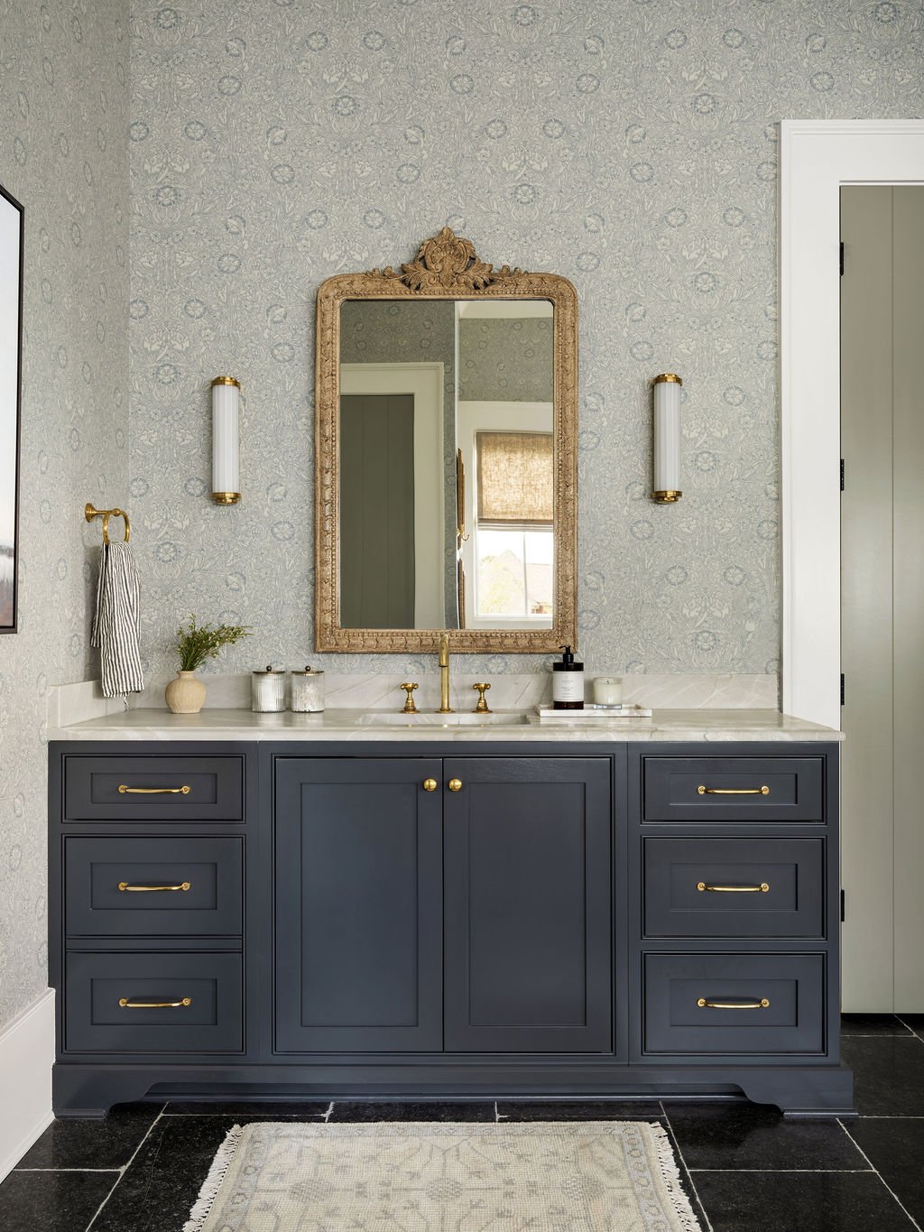 Bathroom vanity with dark gray cabinet, white marble countertop, gold fixtures, and decorative mirror. Items on countertop include soap dispenser, candles, and a small plant. Walls are covered in gray floral wallpaper with gold wall sconces on either