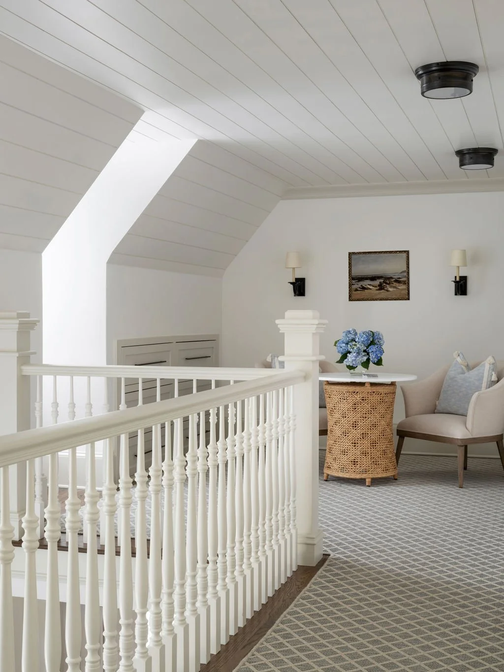A cozy sitting area with white walls and sloped ceiling, featuring two wall-mounted lamps, a framed painting, a wicker side table with blue hydrangea flowers, and two armchairs with cushions, next to a white staircase railing.
