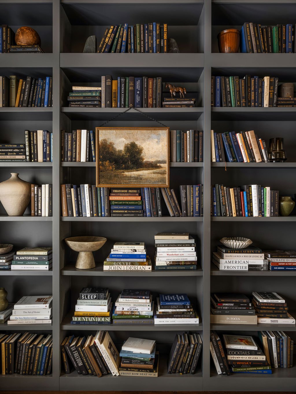 A large gray bookshelf filled with books, decorative objects, and a framed landscape painting hanging in the middle.