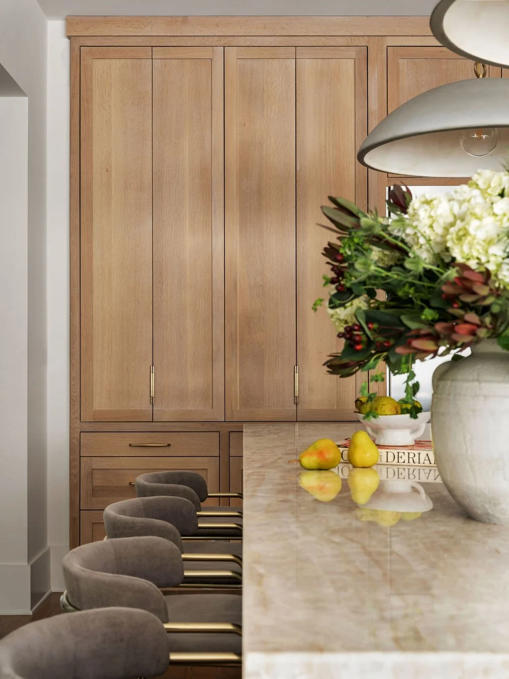 Modern kitchen with beige wooden cabinets, a marble countertop, a large vase with white and dark red flowers, yellow pears, and beige upholstered chairs with brass accents.