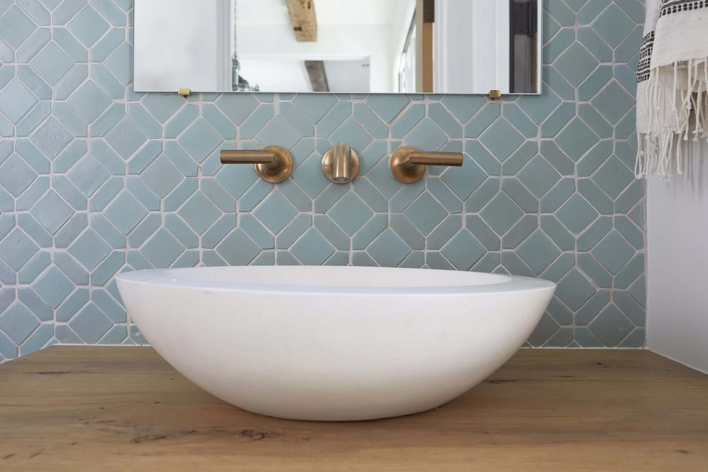 Close-up of a minimalist bathroom sink with a white, rounded basin on a wooden countertop. Behind the sink, a wall with patterned blue tiles features three gold-colored fixtures: two horizontal faucet handles on either side of a central automatic fau