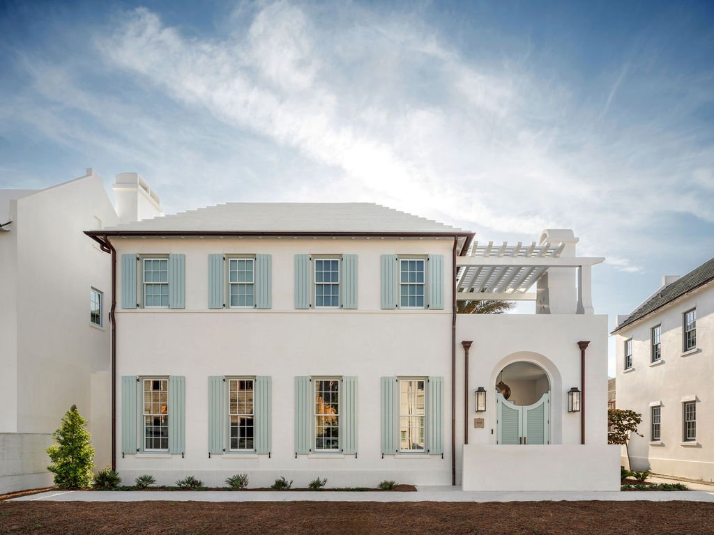 A white two-story house with light blue shutters and a decorative arch above the front door, surrounded by similar styled houses on a sunny day.