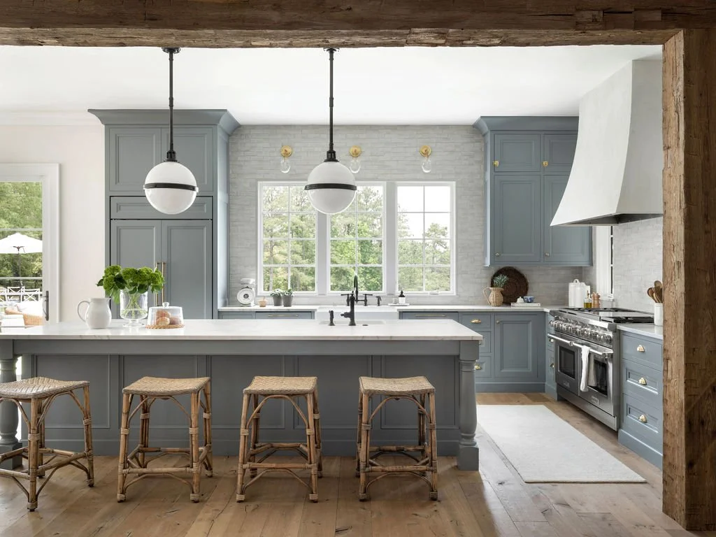 A modern kitchen with blue cabinets, white countertops, a large window, pendant lights, and wooden stools at an island.