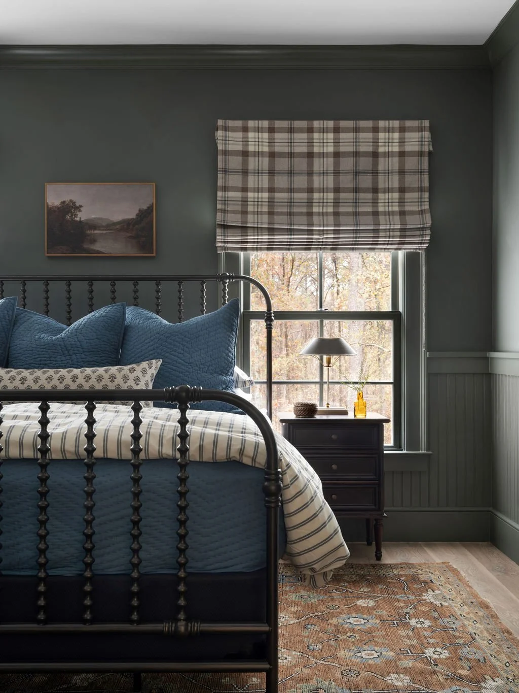 A cozy bedroom with a metal bed frame, blue bedding, a plaid window valance, a small black nightstand with a lamp, a yellow vase with greenery, a picture on the wall, and a patterned rug, all with natural light coming through the window.