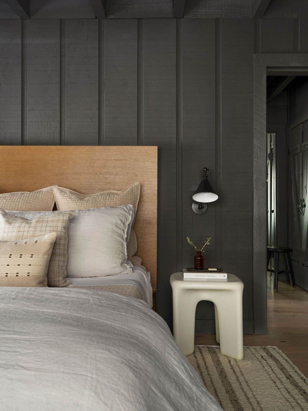 A bedroom with a bed featuring beige and white pillows, dark wooden wall paneling, a white side table with a small brown vase holding a branch, and a black wall-mounted lamp. The room has a rustic, modern aesthetic.