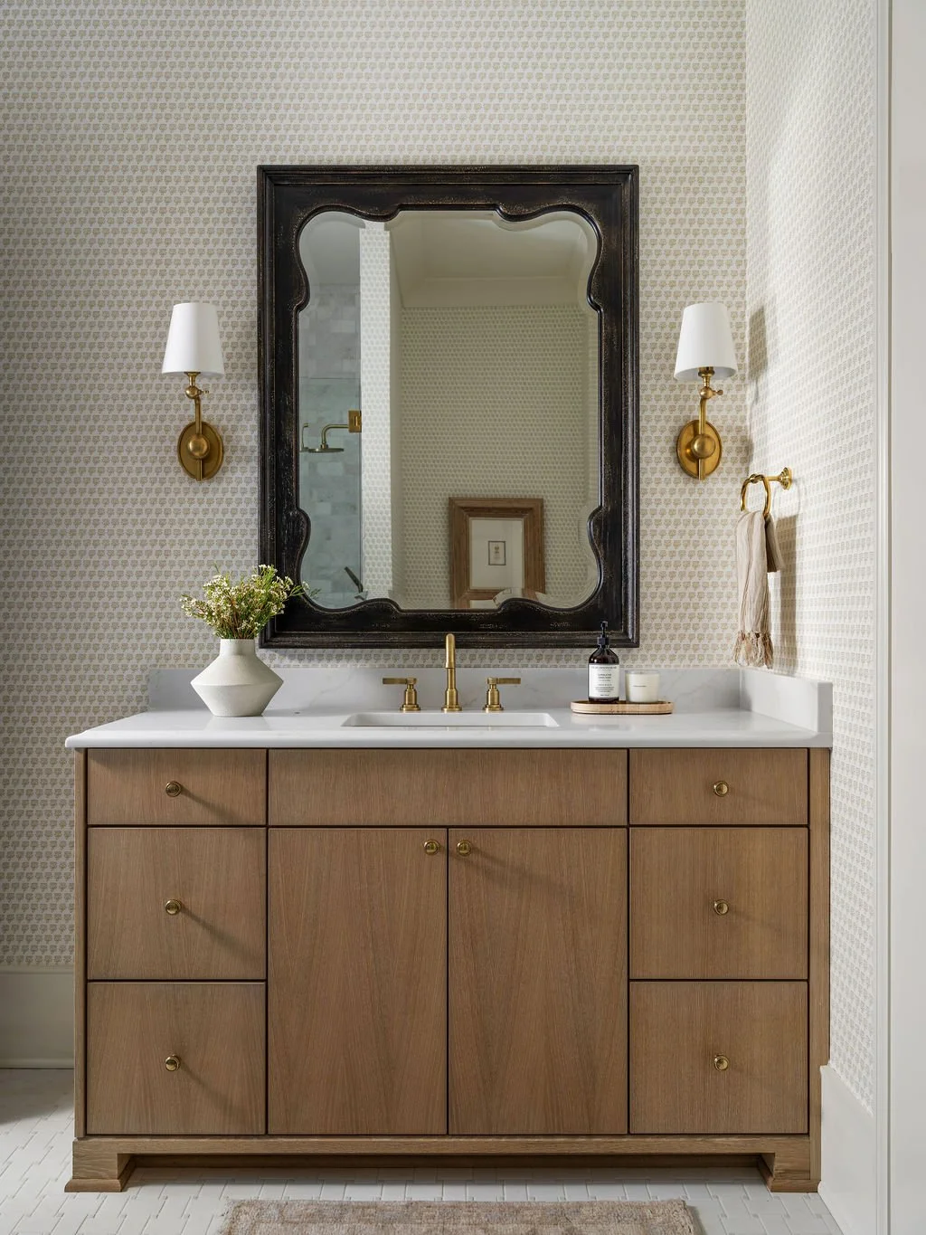 Bathroom vanity with wooden cabinet, white marble countertop, and black framed mirror. Two wall-mounted brass sconces with white shades, a vase with flowers, and toiletry bottles on the counter.