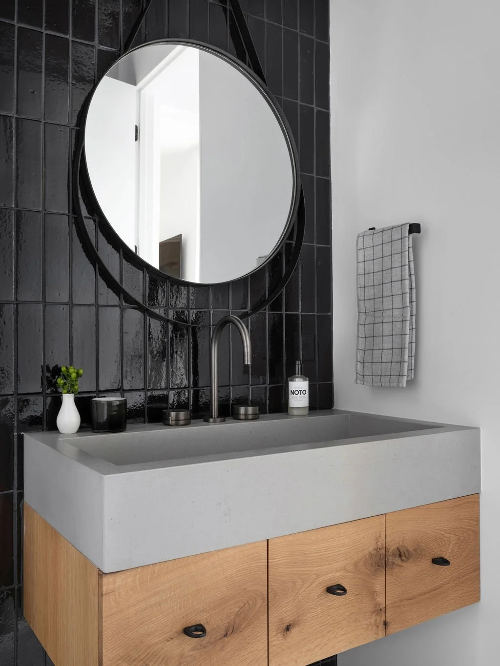 A modern bathroom vanity with a circular mirror, black tiled wall, and a gray sink. The vanity has wooden drawers, a soap dispenser, and small decorative items. A towel hangs on a hook on the wall.