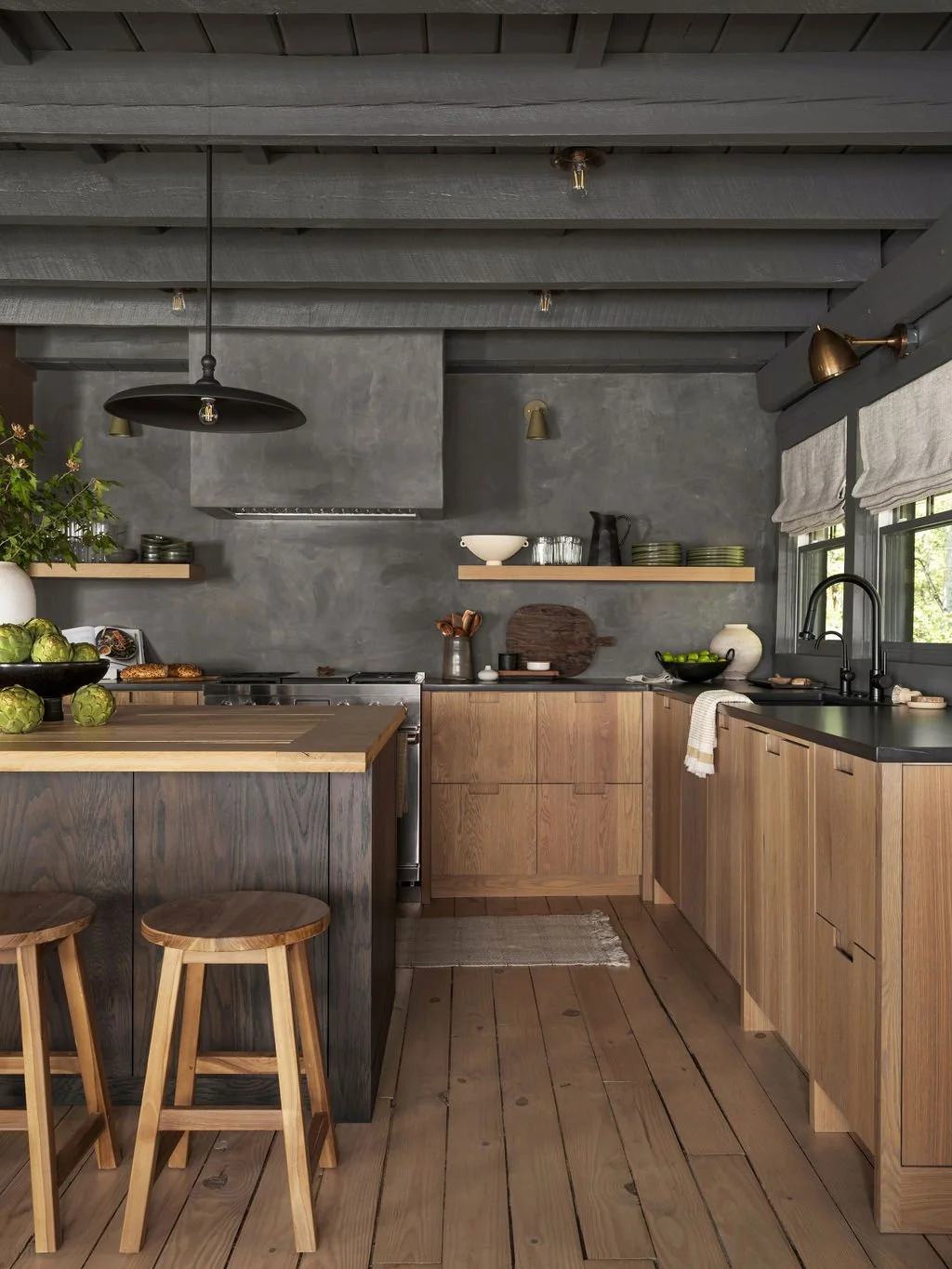 Modern kitchen with wooden cabinets, black countertops, gray concrete walls, hanging black pendant light, and natural wood floor and furniture.
