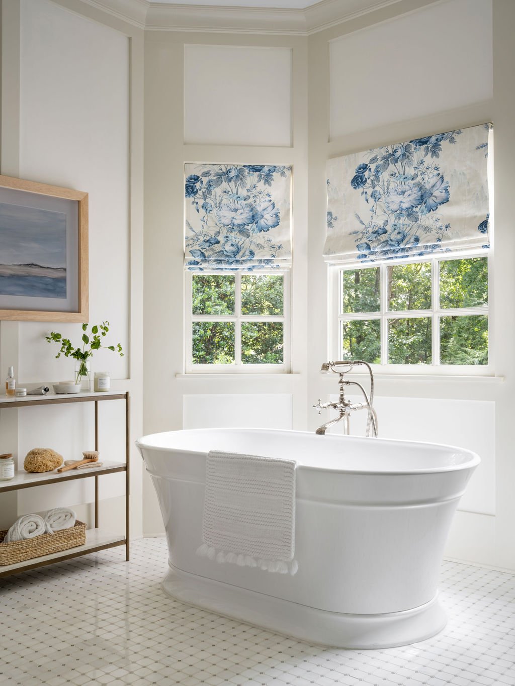A bright bathroom featuring a white freestanding bathtub with a seat towel, a wooden shelf with toiletries, and two windows with blue floral Roman shades.