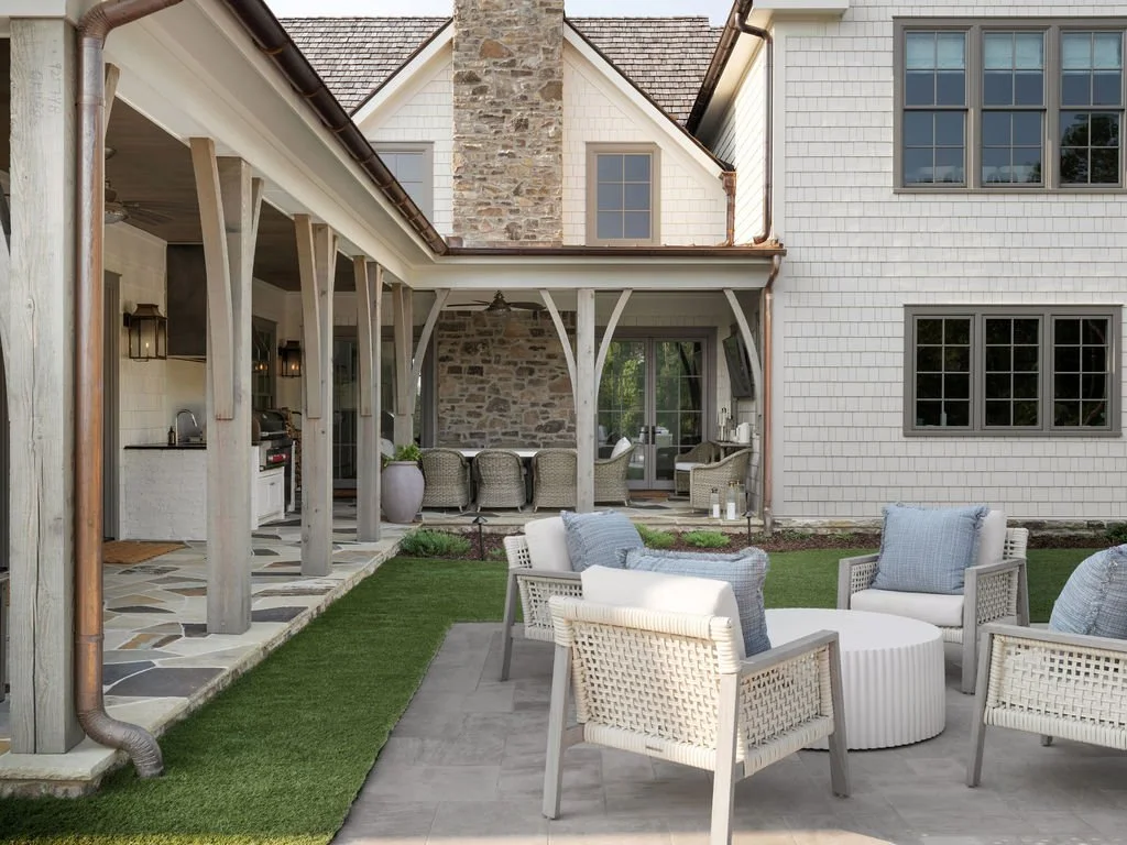 Outdoor patio area with white wicker chairs and a round coffee table, a grassy lawn, and an adjacent covered porch with stone and shingle exterior walls, outdoor kitchen area, and additional seating with view of trees through sliding glass doors.