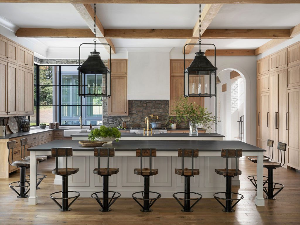 Modern kitchen with a large island, wooden barstools, pendant lights, and a window with greenery outside.
