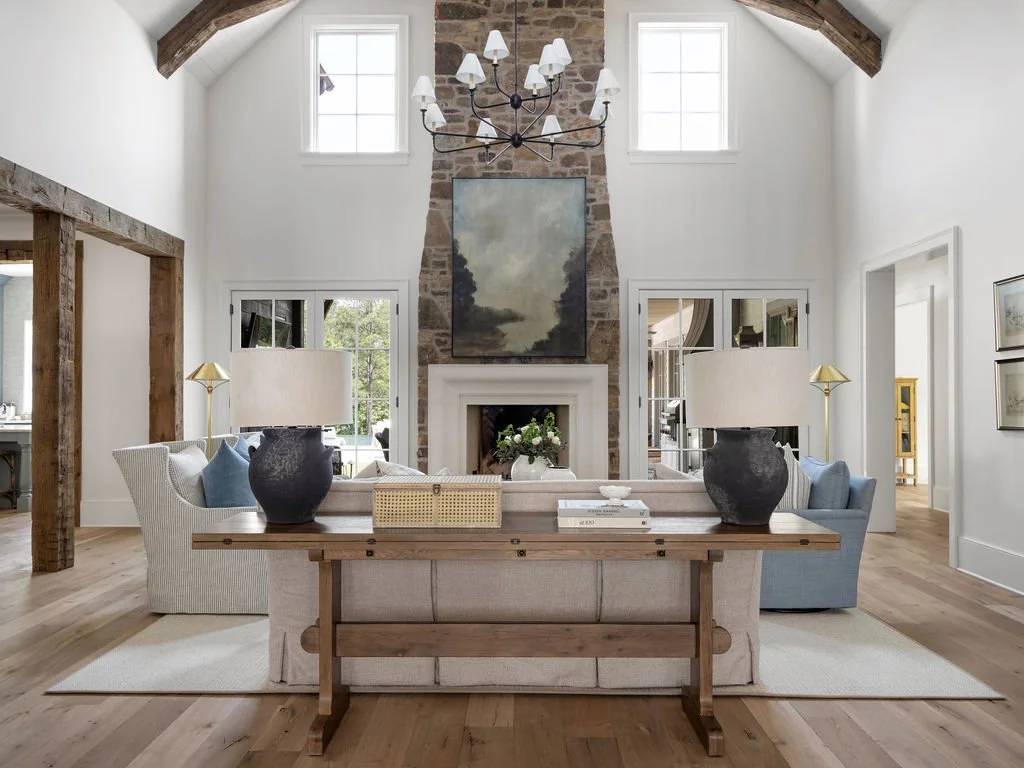 Living room with a high ceiling, exposed wooden beams, a stone fireplace, and a chandelier. White walls and large windows provide natural light. The room is decorated with sofas, lamps, and artwork.