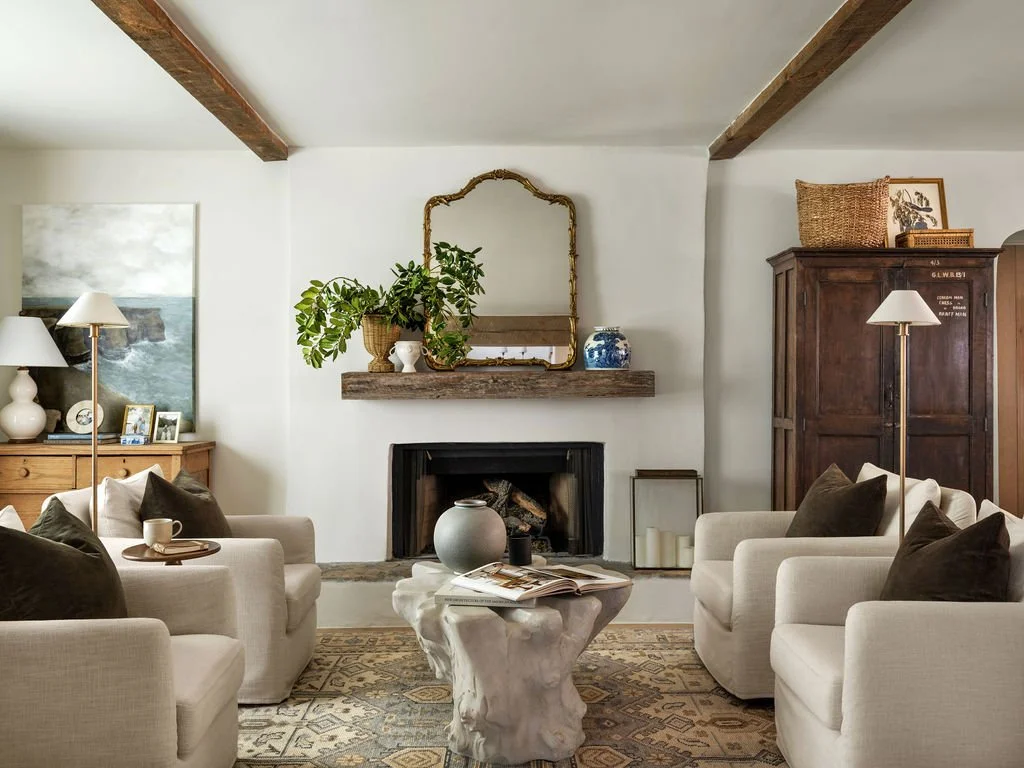 Living room with four beige armchairs, a fireplace with a wooden mantel, a mirror, plant, and decorative items on the mantel, two floor lamps, a wooden cabinet, and a patterned rug.