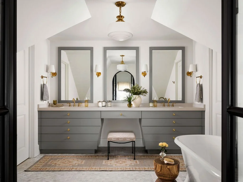 A modern bathroom featuring a double vanity with gray drawers, two mirrors, gold fixtures, and wall sconces. There are two sinks, a central mirror, and a decorative vase with greenery on the countertop. A small stool is placed between the vanity and 