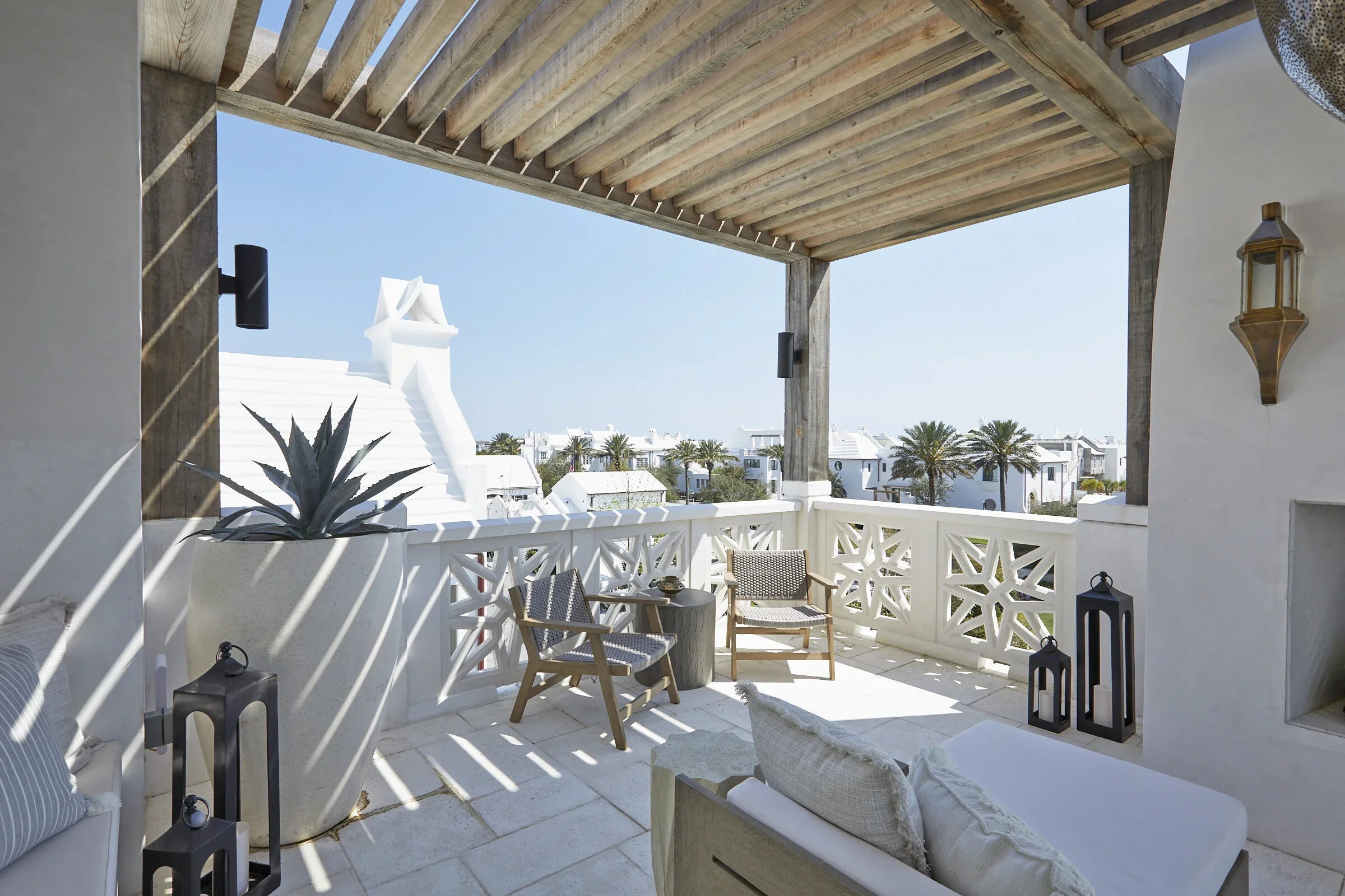 Balcony with white lattice railing, wooden chairs, potted agave plant, and decorative lanterns, overlooking a neighborhood with white buildings and palm trees under a clear blue sky.