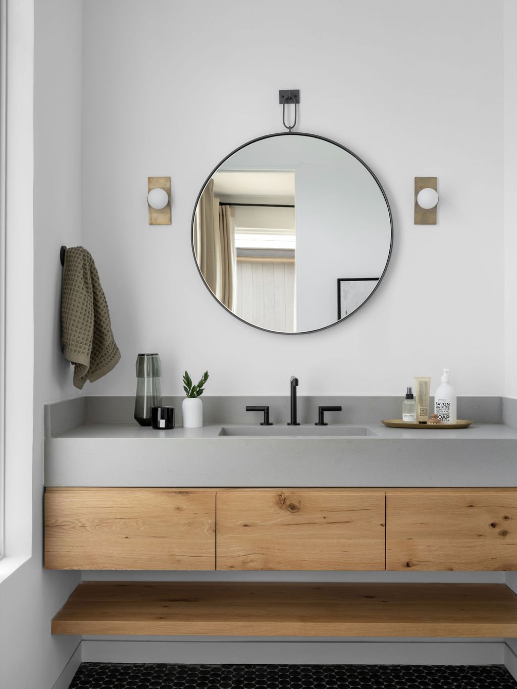 Minimalist bathroom with a round mirror, black wall-mounted faucet, beige towel on the left, and small potted plant on the light gray countertop. Items include soap, lotion, and a tray, with wooden cabinetry below and hexagon black floor tiles.