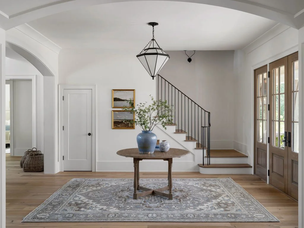 Interior view of a bright entryway with a round wooden table, a large blue vase with greenery, framed landscape art, a staircase with black iron railing, wooden double front doors, and a patterned area rug.