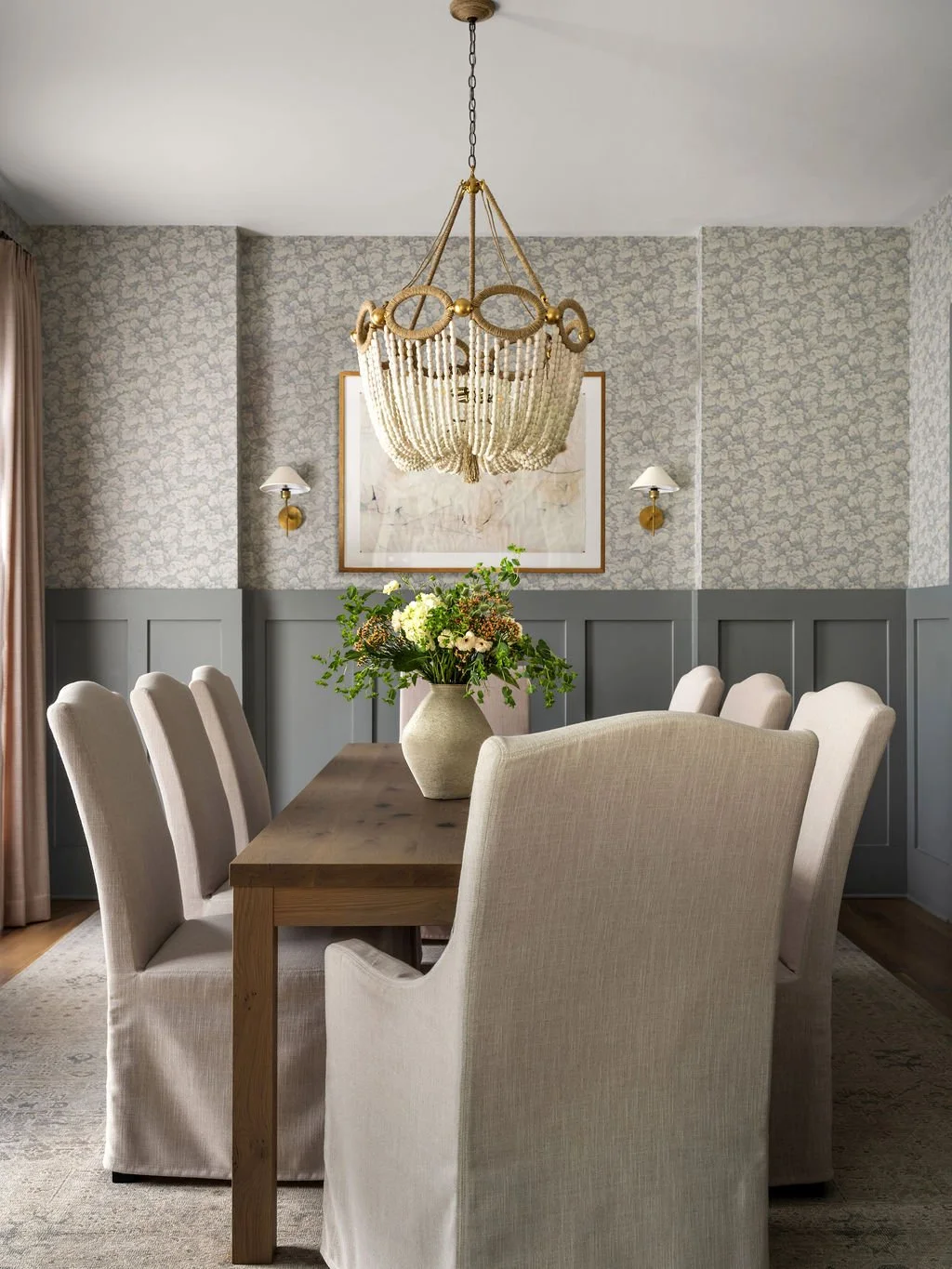 A dining room with a wooden table, cream-colored upholstered chairs, a floral centerpiece in a vase, patterned wallpaper, wall sconces, a framed artwork, and a chandelier.