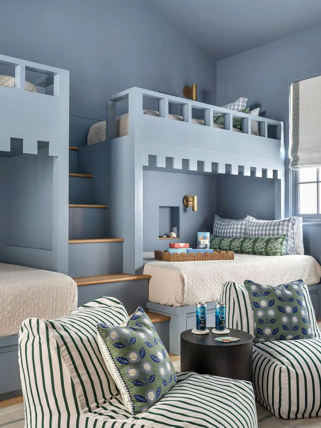 A room with built-in bunk beds painted in light blue, featuring a space for sitting and sleeping, decorated with pillows and blankets. There are striped and floral pillows, a window with curtains, and a small round table with drinks and snacks in the