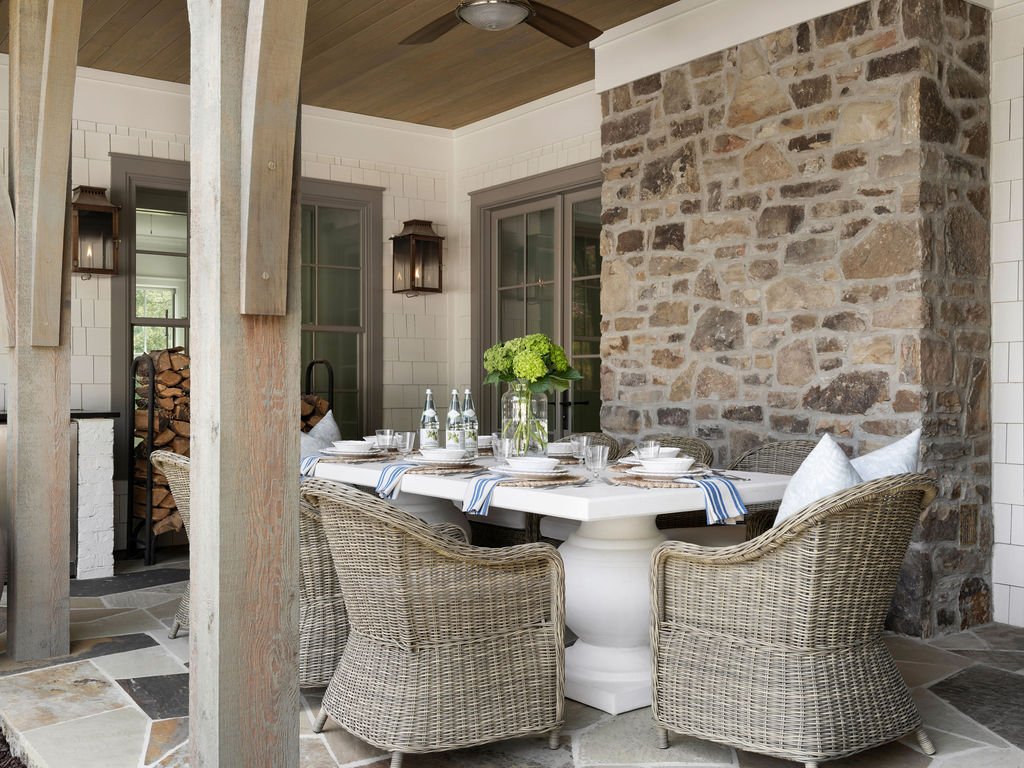 Outdoor dining area with a white table, wicker chairs, a stone fireplace, and large windows with green curtains.