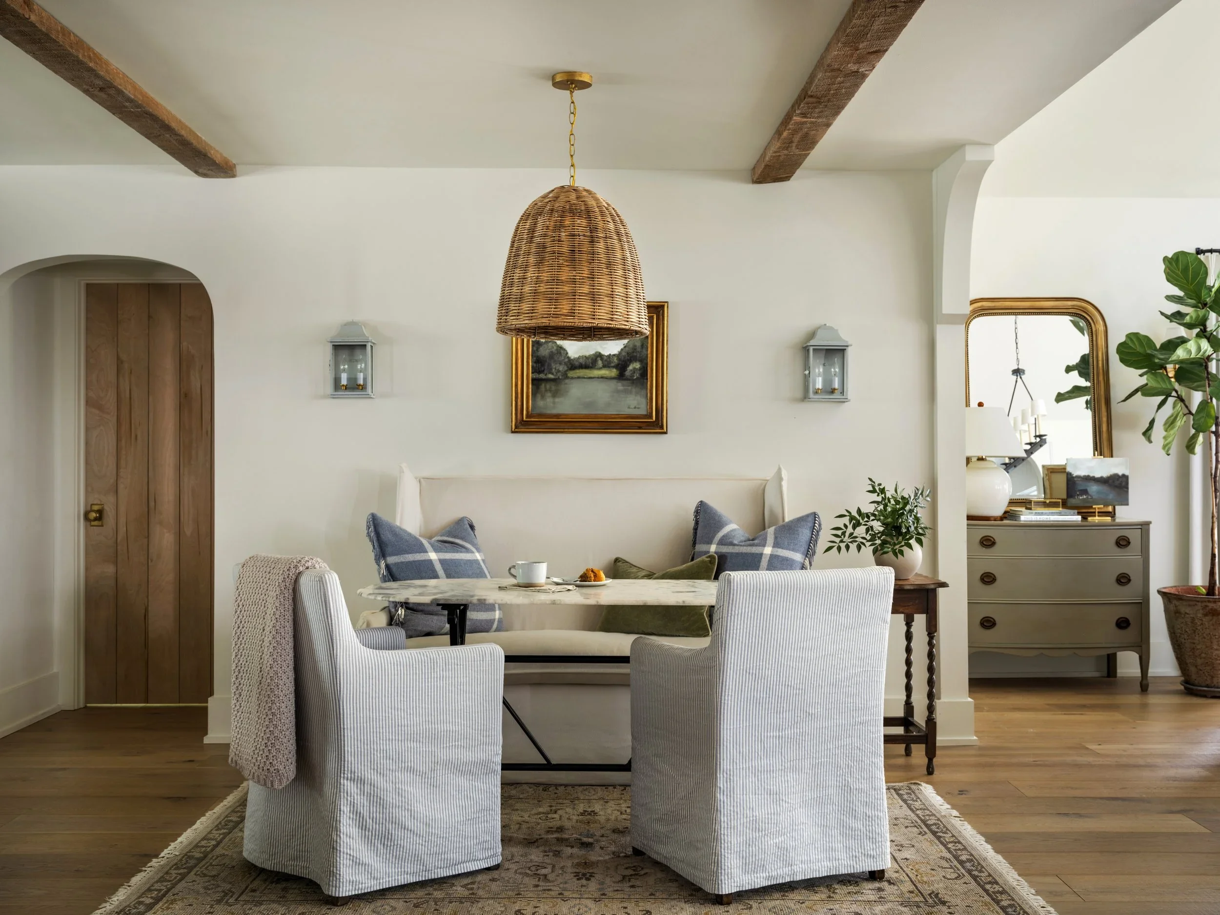 A dining room with a white bench and four striped upholstered chairs around a round marble table. There is a woven pendant light hanging from the ceiling, framed landscape painting, wall-mounted lanterns, a large mirror, and various decorative items,