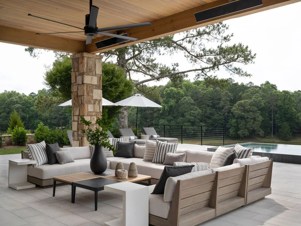 Outdoor patio with white sectional sofa, pillows, a black coffee table, potted plant, sun umbrellas, and a view of a wooded landscape and pool.
