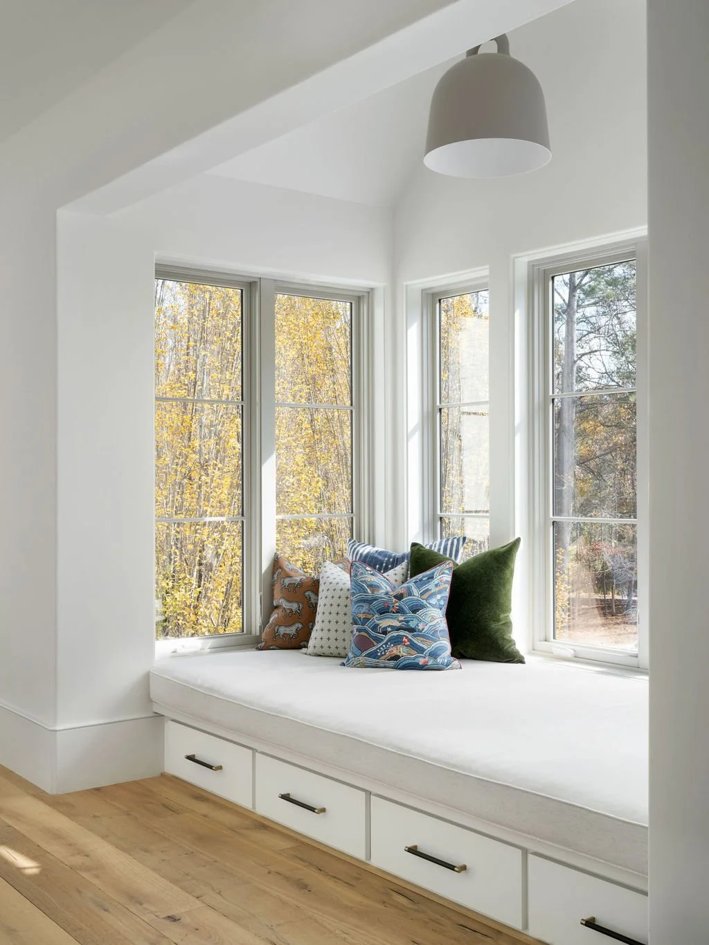 Sunlit window seat with colorful patterned cushions in a bright white corner nook with large windows overlooking trees with fall foliage.