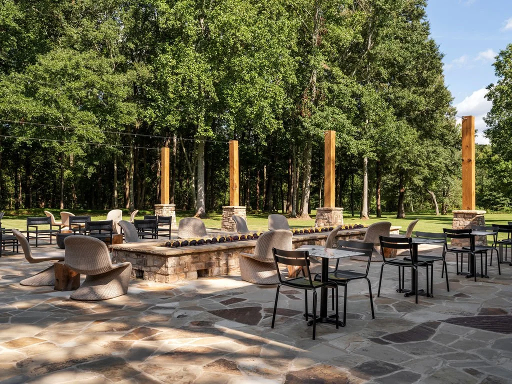 Outdoor patio with stone flooring, several chairs, and a long stone table with a string of small lights on top, surrounded by tall trees.