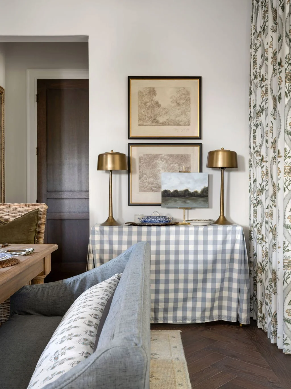 A living room corner with a sideboard covered in blue and white checkered fabric, two gold table lamps, framed artwork, a landscape painting, and curtains with a floral pattern. Part of a gray sofa and a wooden dining table with placemats and a woven chair are visible.