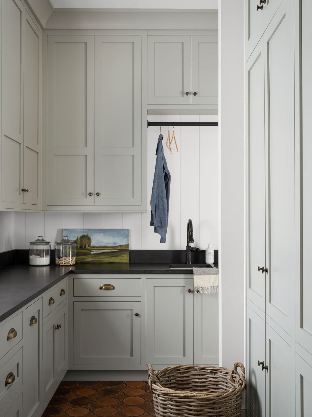 A corner of a kitchen with light gray cabinets, a black countertop, a sink with a black faucet, and a woven laundry basket on a brown patterned tile floor.